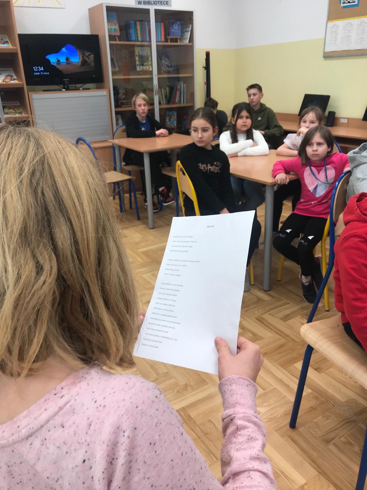 Uczennica czyta poezję Czechowicza na tle słuchających jej uczniów.