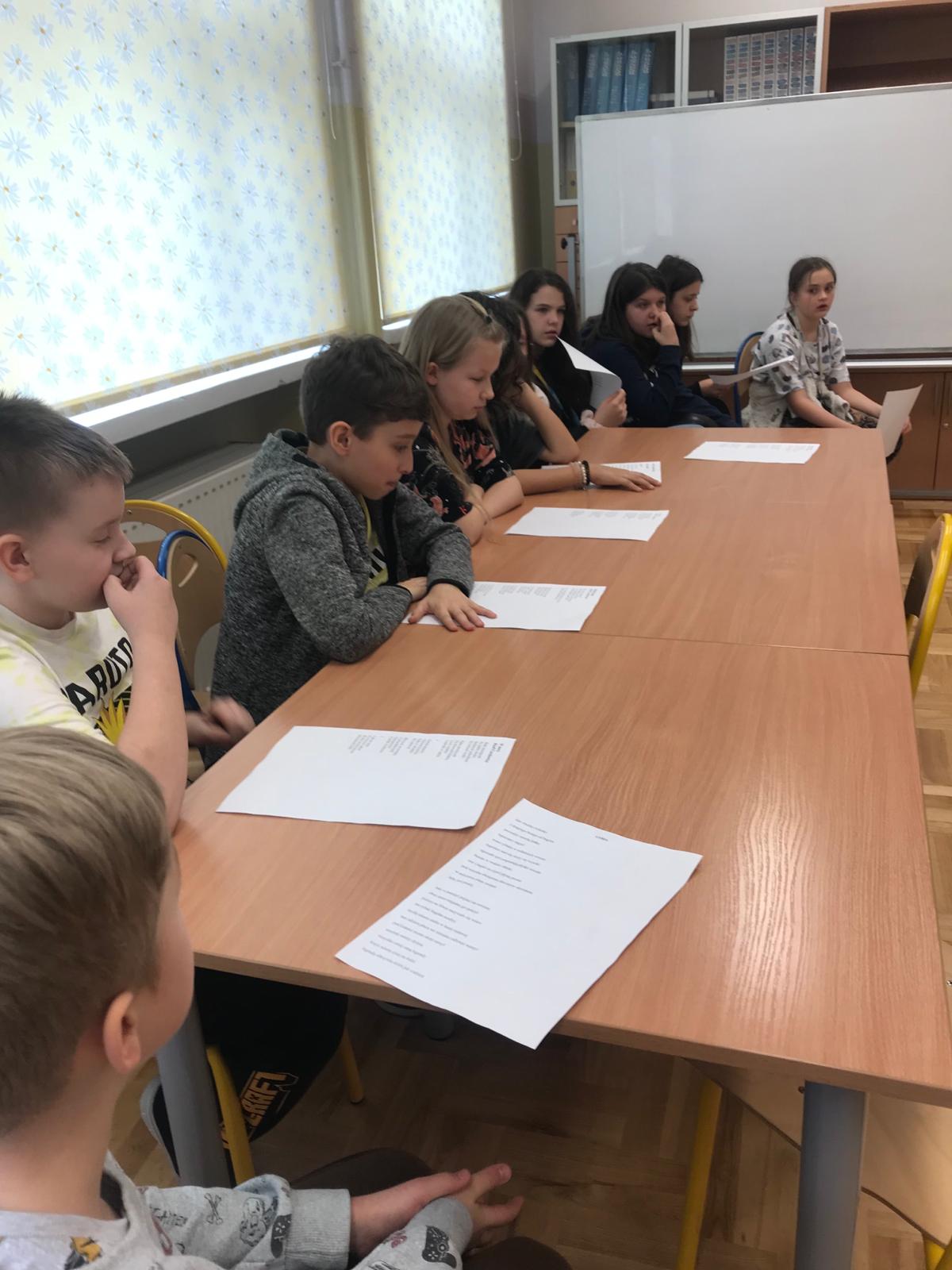 Uczniowie siedzący za stołem czytają poezję Czechowicza.